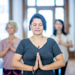 A dark-haired woman sitting in meditation with her hands in prayer position by her heart.