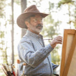 An older man in a cowboy hat, painting at an easel outside.