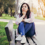 A young dark-haired woman sitting on a park bench, just gazing and thinking.