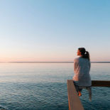 A woman in a ponytail, sitting on a railing, gazing off at the ocean in front of her.