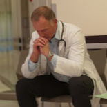 A doctor sitting alone on a chair with his hands clasped under his chin, thinking.