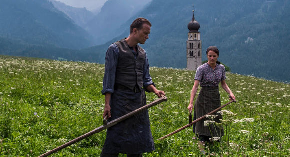 August Diehl as Franz Jagerstatter and Valerie Pachner as Franzisaka in a flower-filled field with mountains in the background.in A Hidden Life 