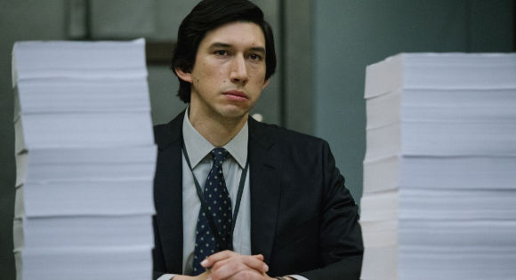 Adam Driver as Daniel Jones in The Report. He is sitting with the report: two gigantice piles of paper on the table to either side of him.